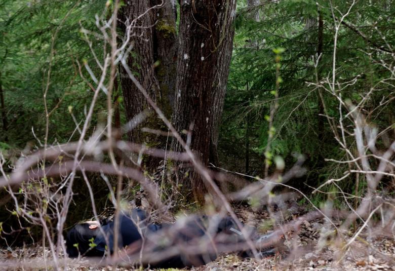 Kuvassa on taustalla vihreää havumetsää ja keskellä on tumma kuusen runko sekä etualalla paljaita oksia.  Maassa näkyy pienesti mustassa asussa käpristynyt ihminen.