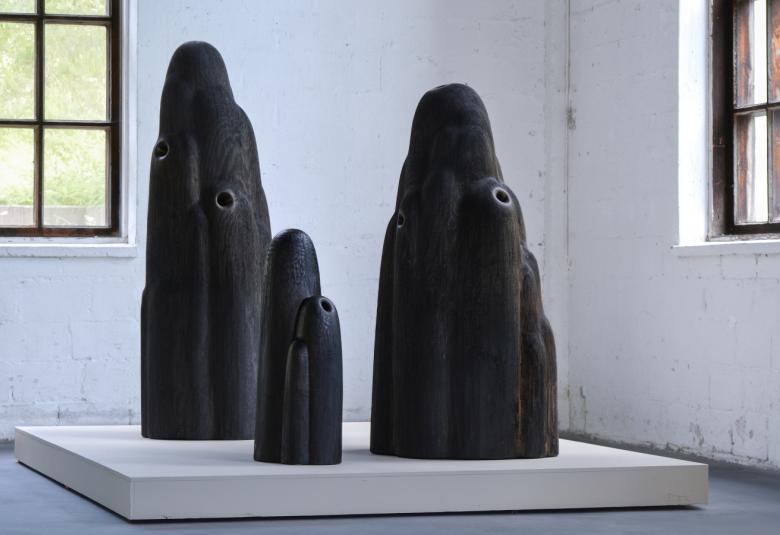 Kuvassa on jalustalla kaksi tummaa eri kokoista patsasta, joista keskellä on pieni.  Teokset ovat huoneen kulmassa ja molemmissa seinissä on ruutuikkunat.