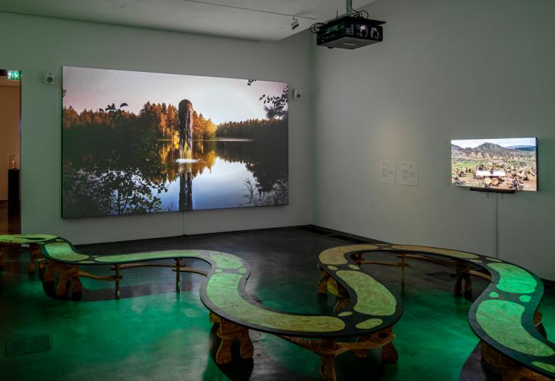 Kuvassa on etualalla vihreä kiemurteleva penkki.  Takana seinällä on taideteoksia, joista takaseinällä on suu tyyntä järven pintaa ja sen takana olevaa metsää esittävä teos ja oikealla pienempi teos.  