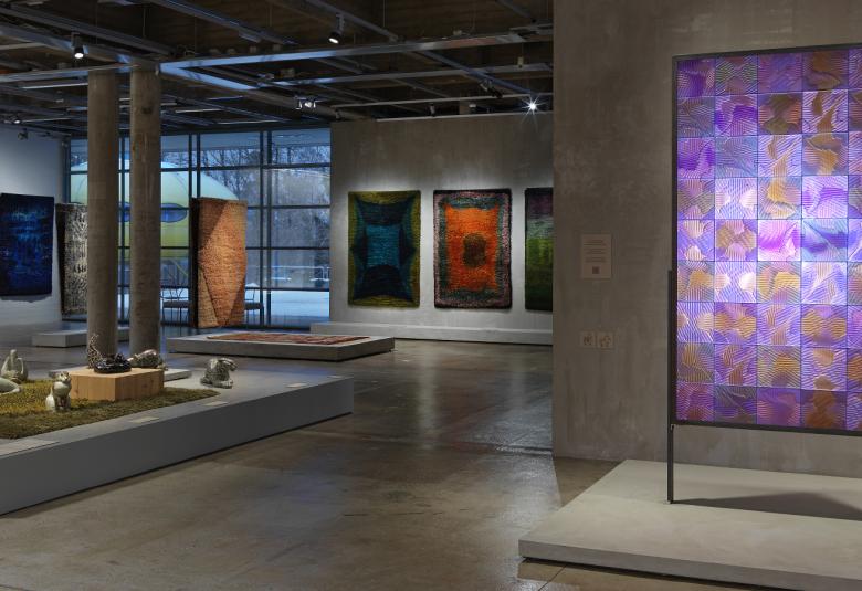 Kuvassa on oikealla etualalla punavoittoinen ryijy ja taustalla useita ryijyjä seinällä. Keskellä jalustalla on keramiikkaa.