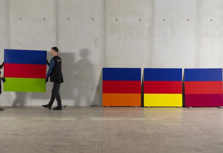 Kuvassa on vasemmalla kaksi henkilöä kantamassa isoa levyä, jossa on tasaraidat sinistä, punaista ja vaalean vihreää.  Vieressä on lattialla nojaamassa seinään vastaavia tauluja, jotka on jaettu tasaisesti sinisellä, punaisella, keltaisella tai tummalla oranssilla. Seinä on harmaa betoniseinä.