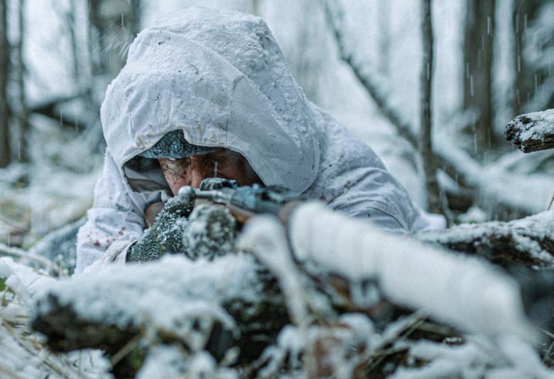 Kuvassa on sotilas valkoisessa lumipuvussa makuulla oksaisessa lumihangessa ampuma-asennossa ase eteenpäin.