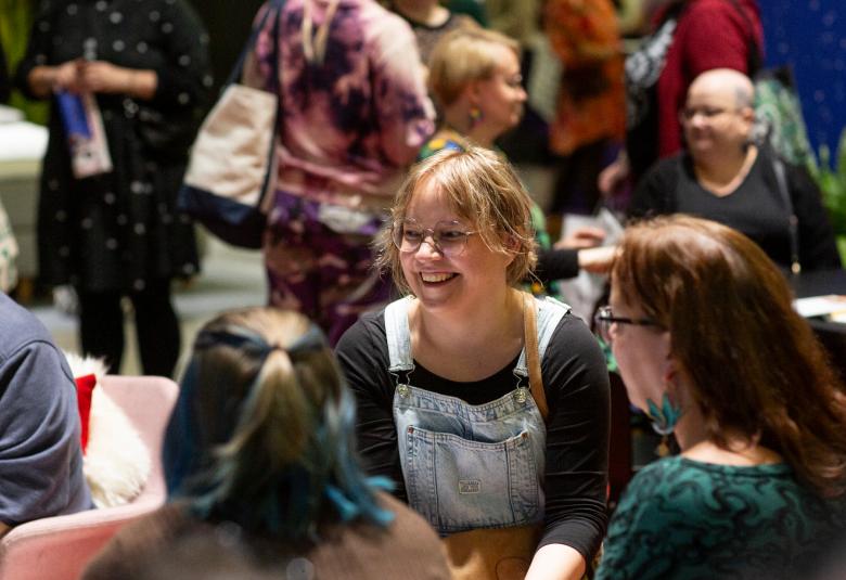 Kuvassa on kirjamessujen yleisöä ja tarkennettuna hymyilevä nuori nainen istumassa ja juttelemassa ringissä olevien toisten naisten kanssa.