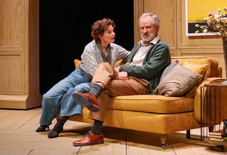 Kuvassa ovat Minna Hämäläinen kääntyneenä Taneli Mäkelän puoleen ja istumassa kellertävällä sohvalla. TAsutalla on ruskeaa puuseinää ja vasemmalla ruskeat puiset kaapin ovet. 