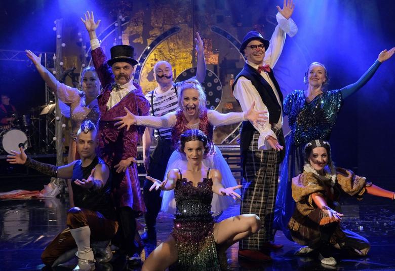 Kuvassa on ryhmäkuva shown esittäjät muodostelma-asetelmassa.  Heillä on reunimmailsilla käsi ylhäällä ja siitä tulee kaarimainen asetelma.  Heillä on show-vaatteet päällään.  Tausta on tumma.