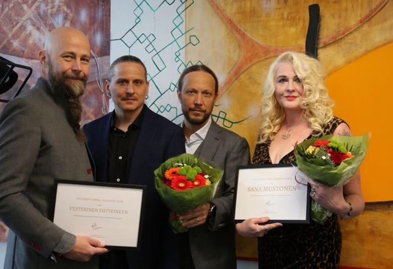 Kuvassa on vasemmalla kolme miestä, joista oikean puoleinen Tero Vesterinen pitää kädessään kehystettyä kunniakirjaa.  Oikealla on Sana Mustonen, jolla on vaaleat, puolipitkät hiukset ja hän pitää kädessään kunniakirjaa. 