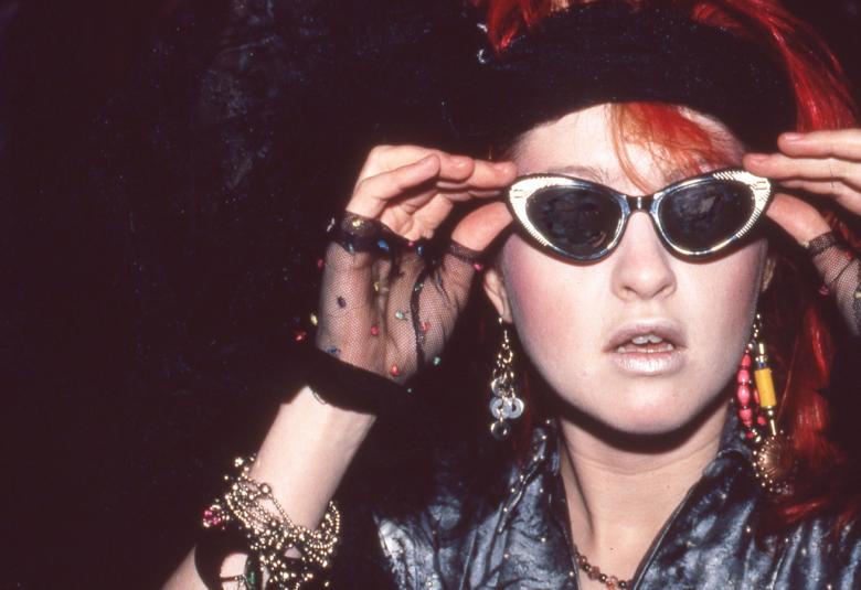 Kuvassa ovat nuoren naisen kasvot ja hänellä on aurinkolasit ja pitää sangoista kiinni molemmilla käsillään. Hänellä on päässään musta hattu ja tausta on tumma.