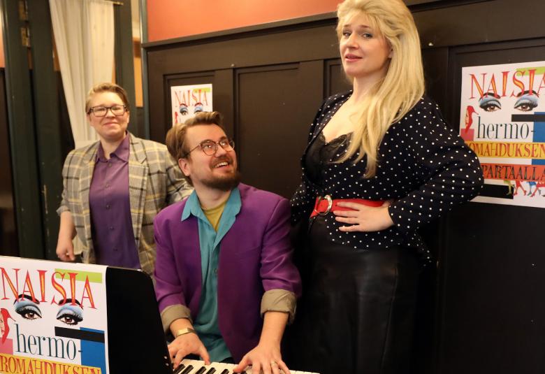 Kuvassa on taaempana Eve Pietarinen ja edessä Kasper Omenasaari sähköpianon ääressä.  Hän katsoo seisomassa olevaa Regina Launivuota.  Takana on ruskeaa puuseinää, joka on puolet seinästä.  Edessä on näytelmän juliste pöydällä pienikokoisena ja kaksi seinällä.