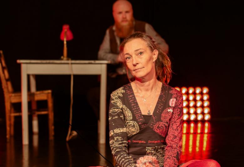 Kuvassa on etualalla nainen istumassa lattialla ja hänellä on ruskean kirjava mekko. Takana istuu mies ja hänestä näkyy pää. Taustalla on vaalean sininen pöytä ja ruskea tuoli. 