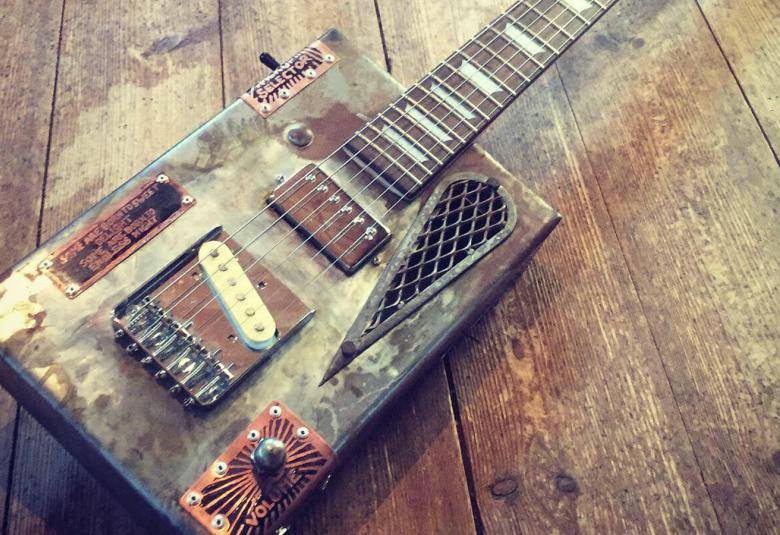 Kuvassa on sikarilaatikko kitaran osana ja siinä on tekstiä ja se on ruskehtava. Laatikosta lähtee kitaran kaula. Taustana on ruskehtava lattia.  