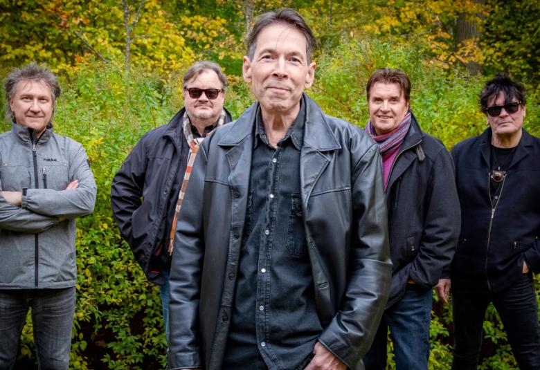 Kuvassa on yhtyeen viisi soittajaa seisomassa ulkona vihreää heinikkoa ja kumpua taustalla. Heillä on tummat pusakat päällään.