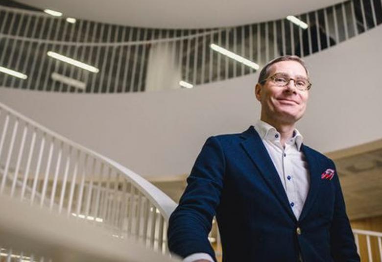 Kuvassa on Tuomas Heikkilä kuvattuna alhaalta päin ja hän seisoo vaaleassa porraskäytävätilassa.  Tila on vaalea ja siinä näkyy kiertävää valkoista kaidetta, jossa on valkoinen pystyraudoitus.