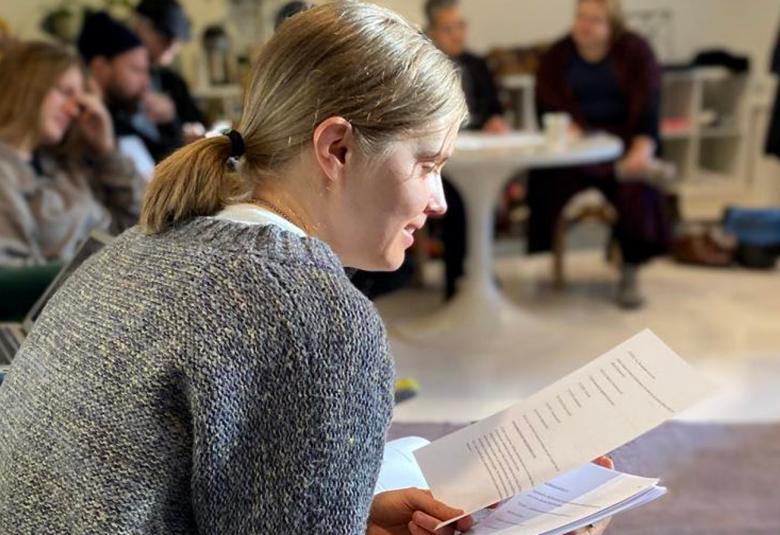 Kuvassa on edessä sivuttain Alina Tomnakov istumassa ja kädessään tekstiliuskoja. Vierestä lähtee näyttelijöiden rivi ja he kaikki ovat istumassa sekä lukemassa  lukemassa paperiltaan.