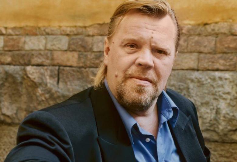 Kuvassa on Vesku Loiri puolivartalokuvassa. Hänellä on tumman sininen takki ja vaalean sininen kauluspaita päällään.  Taustalla on vaalean ruskeaa tiiliseinää. 