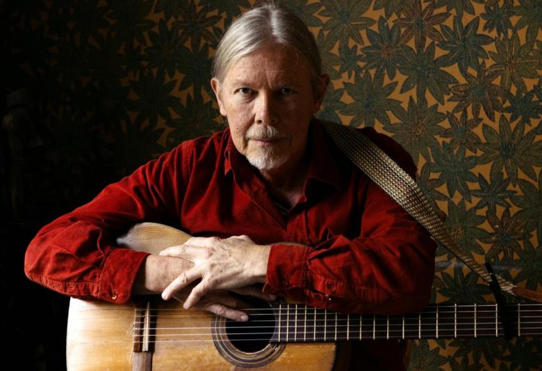 Kuvassa on J. Karjalainen on istumassa kitara sylissään. Hänellä on punaruskea paita päällään ja taustalla on tummahko kukallinen tapetti oikealla puolella kuvaa.