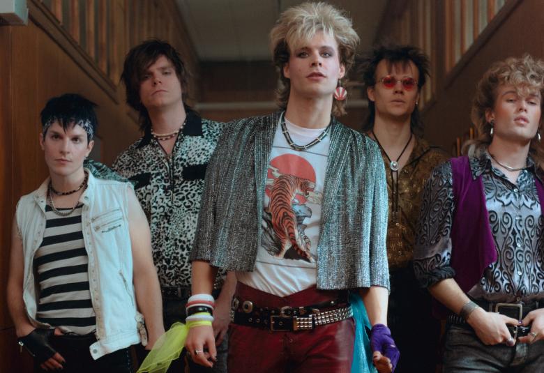 Kuvassa on keskellä Nipa Neumannia esittävä näyttelijä vaaleissa lyhyissä hiuksissa ja hopean harmaassa jakussa sekä kuvallisessa t-paidassa. Vasemmalla ja oikealla puolella on kaksi bändin jäsentä.