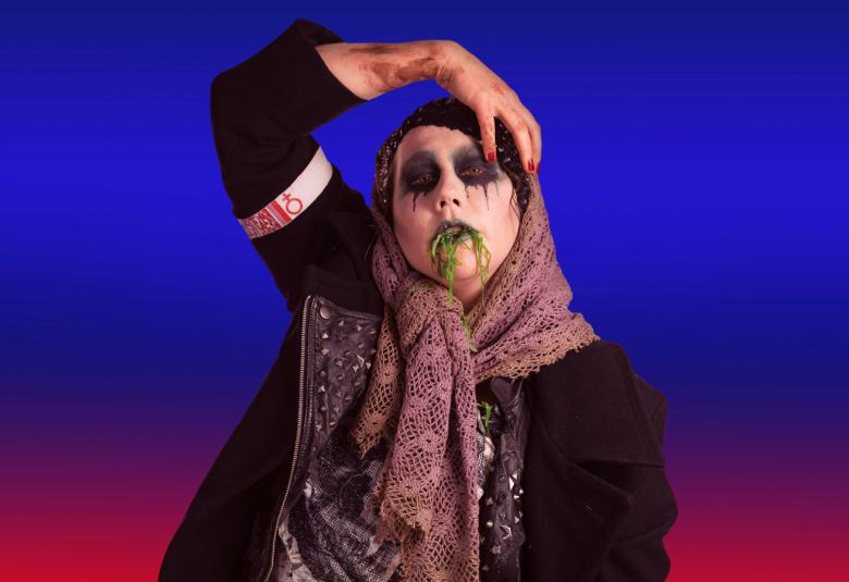 Kuvassa on Annika Aapalahti, joka on pukeutunut isoon tummaan talvitakkiin ja violetin kirjavaan huiviin. Hänen silmänsä on maalattu mustiksi rinkuloiksi ja suupielistä tulee tummat viivat alaspäin.  Hän pitää oikeaa kättään otsallaan. Tausta on violetin punainen.