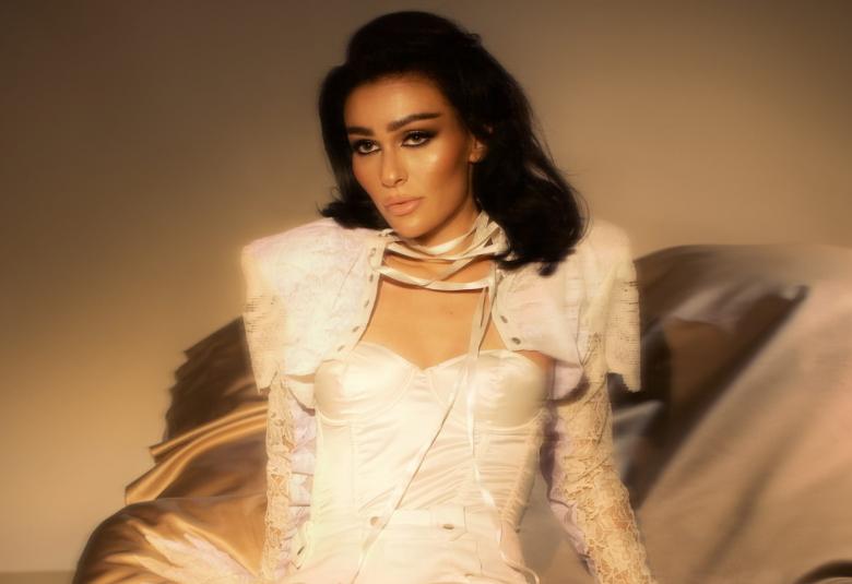 Kuvassa on Sara Siipola istumassa puolivartalokuvassa. Hänellä on valkoinen tiukka asu päällään ja valkoinen, jossa on topatut olkapäät. Tausta on ruskea.