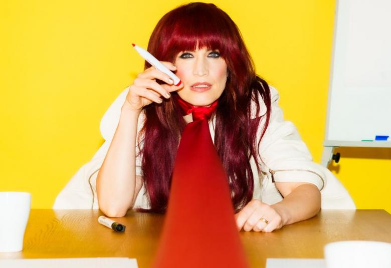 Kuvassa on Maija Vilkkumaa istumassa pöydän takana valkoisessa puserossa ja isossa punaisessa kraatissa, joka menee pöytää pitkin. Pöydällä on valkoisia papereita ja muki. Seinä on keltainen.