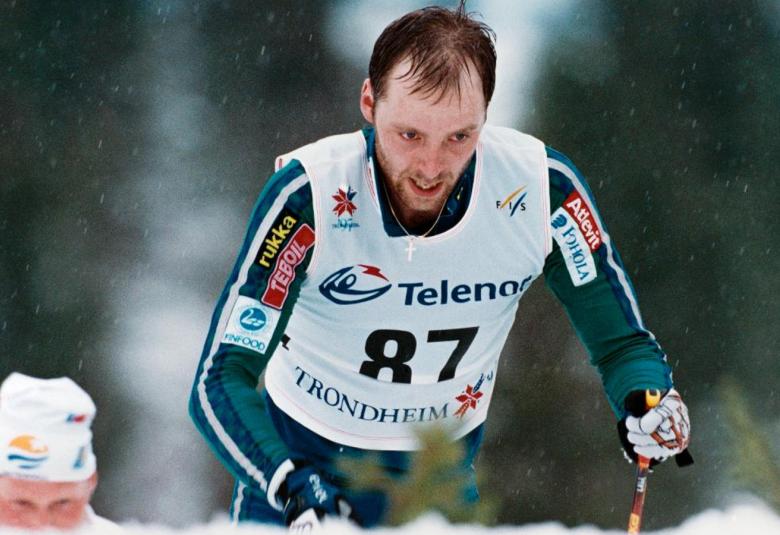 Kuvassa on etualalla Mika Myllylä hiihtämässä sinivalkoisessa hiihtoasussa.  Tausta on harmaa.  Vasemmalla alhaalla näkyy toisen hiihtäjän pää.