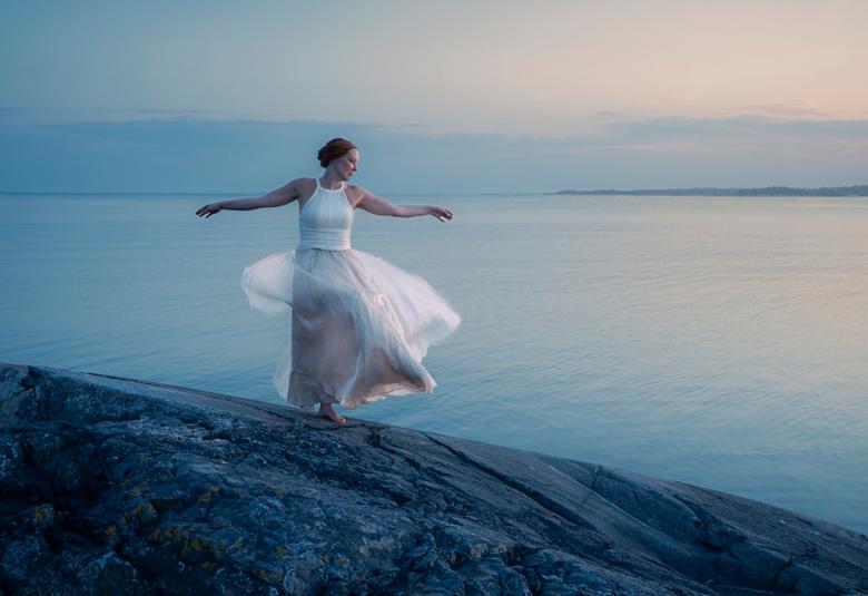 Kuvassa on kalliolla valkoisessa, pitkässä valkoisessa tyllihameessa ja valkoisessa puserossa tanssimassa. Taustalla on sinistä merta taivasta.