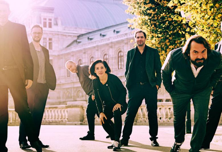 Kuvassa ovat Platform K+K:n soittajat ja he ovat aurinkoisella kadulla. Vasemmalla on mies seisomassa auringon heijastuksessa. Muut joko seisovat tai ovat kumartuneena ja oikealla oleva mies on kumartuneena vahvasti eteenpäin. Taustalla on vaalea 1800-luvun rakennus. 