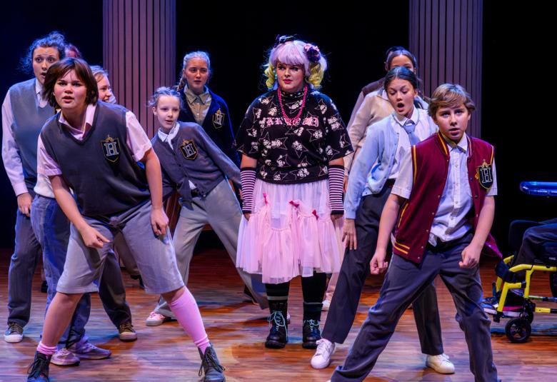 Kuvassa on joukko nuoria tanssimassa siisteissä pitkissä housuissa ja koululiivissä.  Keskellä on tyttö, jolla on vaalea tyllihame ja kirjava pusero sekä vaaleat hiukset. 