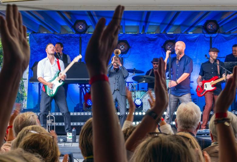 Kuvassa on lavassa kesällä Vesterinen yhteineen soittamassa 10 hengen bändinä. Lavan tausta on kirkkaan sininen. Etualalla on ylös nostettuja käsiä.