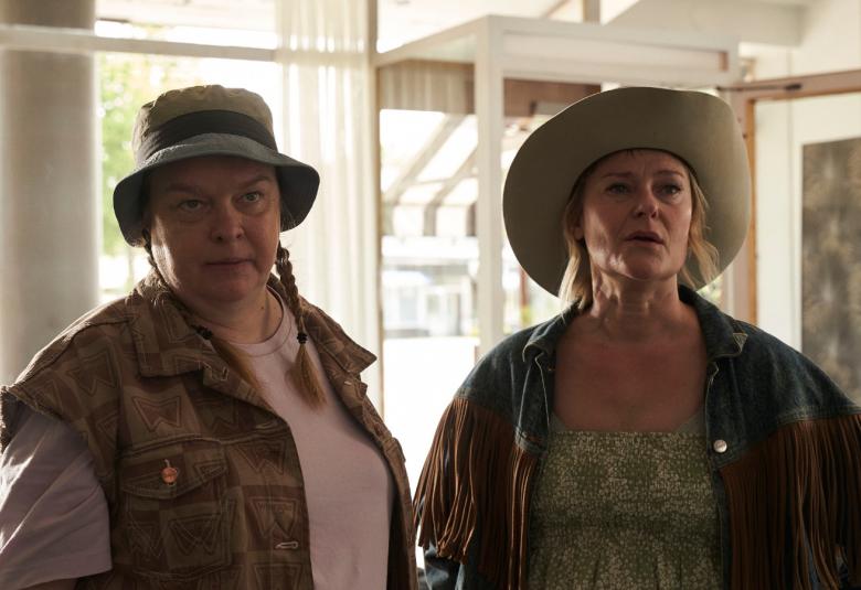 Kuvassa on vasemmalla seisomassa Elina Knihtilä päällään vihertävä liivi. Oikealla on PIrjo Lonka farkkutakissa ja lierihatussa. Molemmat seisovat ja kuva on puolivartalokuva.