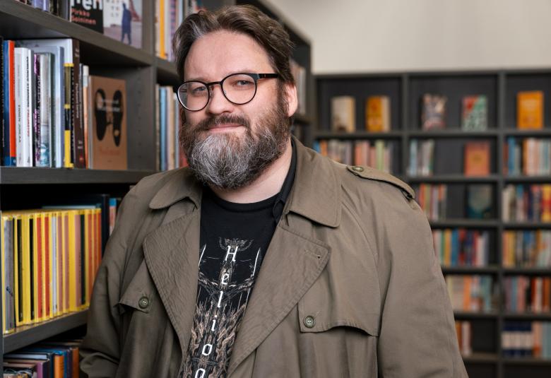 Kuvassa on Ville Hänninen seisomassa puolivartalokuvassa vihertävä trenssitakki päällään.  Hänellä on partaa ja tummasankaiset silmälasit päässään. Taustalla näkyy kirjahyllyä. 