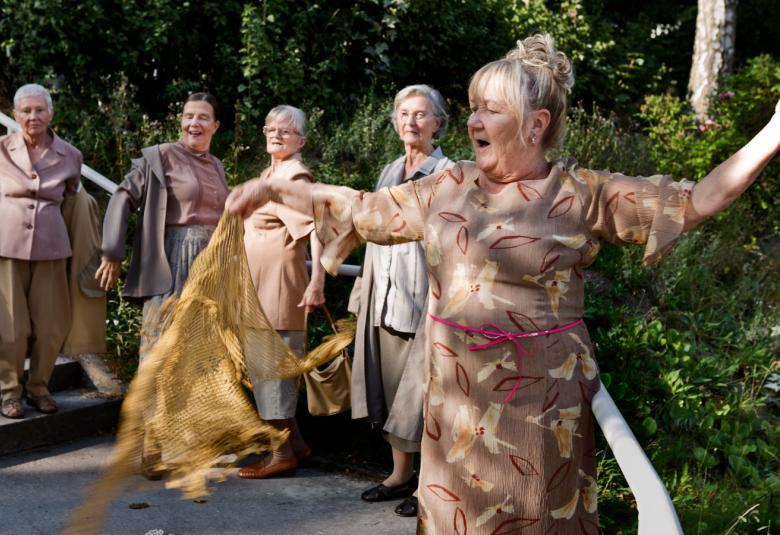 Kuvassa on viisi naista rivissä vasemmalta kiertyvässä jonossa ja riisuvat harmahtavia jakkujaan. Tilalle näkyvät mekot.  Taustalla on vihreää puistometsää.