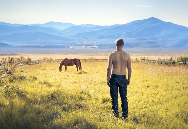 Kuvassa on vihertävän keltaista ruohopeltoa sängellä ja edessä oikealla on seisomassa  mies selin ja ilman paitaa. Kaempana pellolla näkyy hevonen.  Taustalla on vuoria.