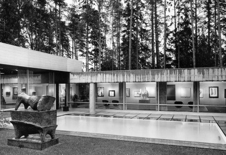 Kuvassa on Didrichsenin taidemuseeon rakennus ja kuva on musta-valkoinen.  Rakennus on matala ja se tekee kulman ja siinä on isot ikkunat. Edessä on patsas, jossa on kivinen jalusta ja sen päällä on kolme kiveä. Rakennuksen edessä on vesiallas.