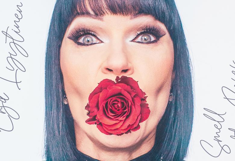 Kuvassa on Erja Lyytinen kasvokuvassa, jossa hänellä on ruusu suussaan. Tausta on vaalea ja vasemmalla on hänen nimikirjoituksensa.  Oikealla on käsialalla Smell the Roses.