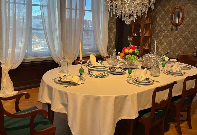 Kuvassa on valkoisellaliinalla katettu ruokapöytä ja kalusteet ovat vanhanaikaisen ruskeita. Huoneessa on korkeat ikkunat ja laskeutuvat vaaleat verhot sekä kristallikruunu.