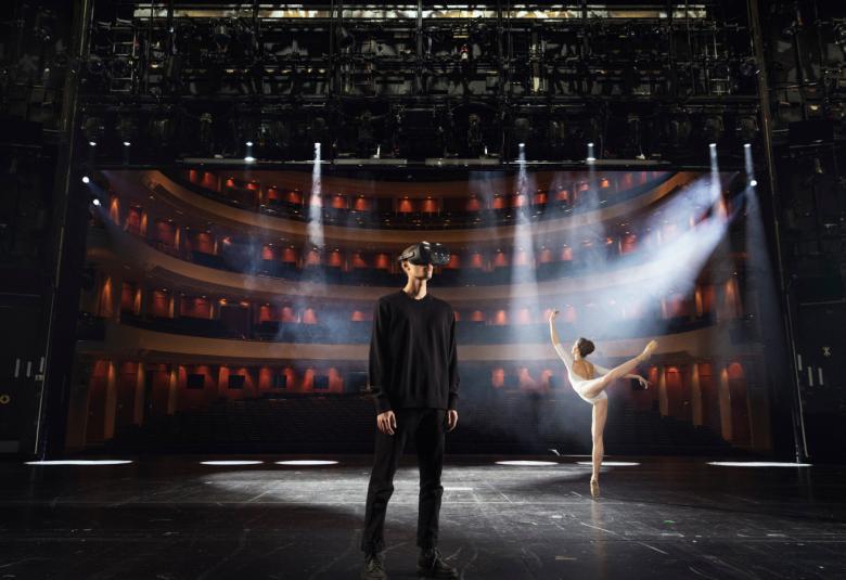 Kuvassa on tummalla näyttämöllä seisomassa mies, jolla on virtuaalilasit päässään. Taustalla näkyy balettitanssija valkoisessa balleriinapuvussa selin ja oikea jalka ja käsi ojentuneinna.