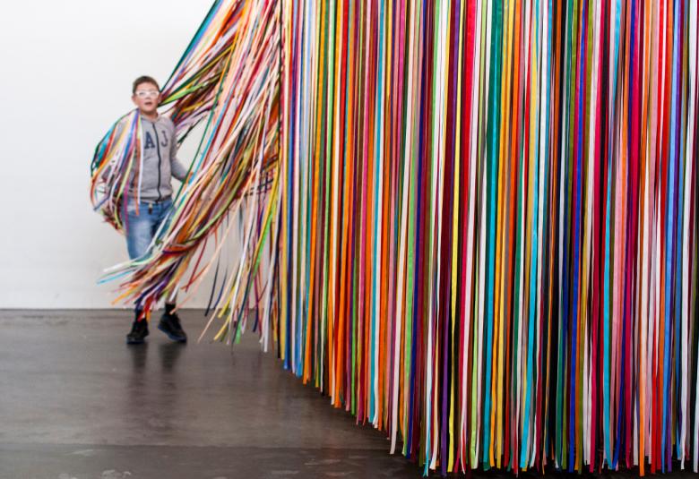 Kuvassa on oikealla eri värisiä ylhäältä tulevia verhonauhoja paksuna seinämänä. Takana on poika, joka on kietoutunut verhonauhoihin. Vasemmalla näkyy vaaleata seinää.