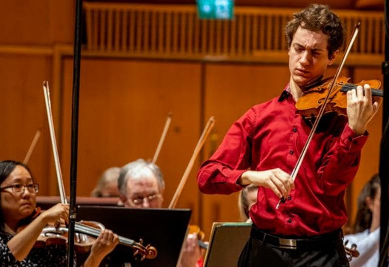 Kuvassa on oikealla Adrián Ibáñez-Resjan soittamassa viulua. Kuva on puolivartalokuva ja hänellä on puna-ruskea paita päällään.  Alempana on orkesteri soittamassa.