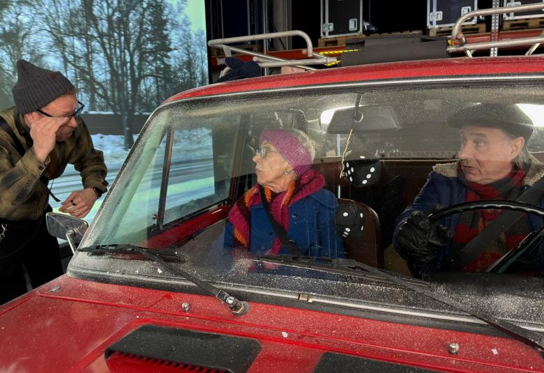  Kuvassa on vasemmalla mies kuvartuneena punaisen auton ikkunan luona ja sisällä ovat Tuija Ernamo ja Tom Lindholm, jotka katsovat häntä. Takana on talvista maisemaa.