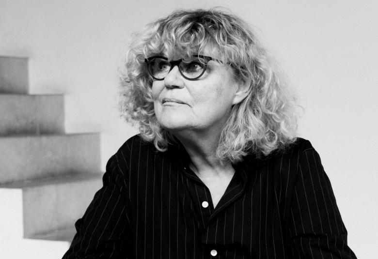 Kuvassa on Marie-José Van Hee istumassa pöydän ääressä. Taustalla on vasemmalla takana valkoiset betoniportaat ja valkoista seinää. Kuva on musta-valkoinen. 