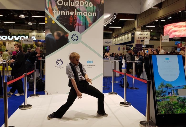 Kuvassa on ilmakitaraa soittamassa Aapo Rautio keskellä olevalla vaalealla esiintymisaleella. Ylhäällä lukee Oulu2026 tunnelmaan. Taustalla näkyy muita esittelyständejä. 