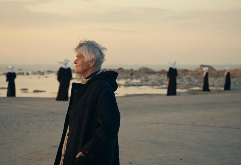 Kuvassa on Pirkko Saisio tummassa päällystakissa hiekkarannalla. Hän katselee edessään olevalle merelle.  Rannalla on tummissa kaavuissa ja valkoisissa myssyissä seisovia naisia pitkin rantaa.