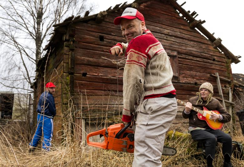 Kuvassa on keskellä ruskea hirsinen riihi ja pitkäruohoista maalaispihaa. Etualalla seisoo mies, jolla on Jussi-paita päällään ja vaaleat housut.  Hänellä on oranssi moottorisaha kädessään.  Hänen takanaan on mies soittamassa mandoliinia kannon päällä. Riihen vieres on selin mies sinisessä verkkarissa.