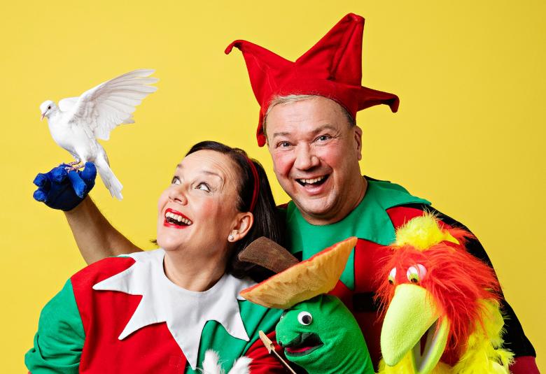 Kuvassa ovat Kirsti Aalto ja Simo Aalto puolivartalokuvassa. Heillä on vihreän punaiset asut ja Simolla punainen monikulmainen hattu päässään. Oikeassa kädessään hänellä on valkoinen kyyhky ja oikeassa kelta-punainen kankainen papukaija.  