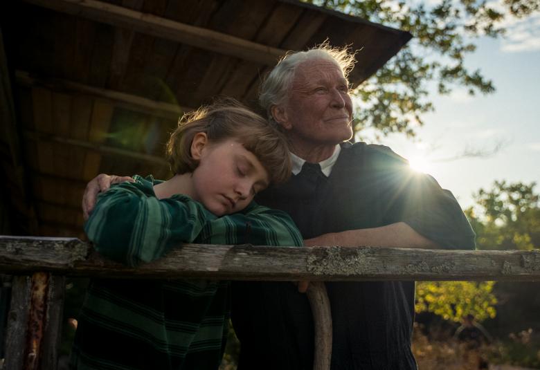 Kuvassa ovat Emily Matthews ja Glenn Close nojaamassa kesämökin kaiteeseen ja he katsovat kauemmas. Taustalla näkyy kesämökin etuseinää. Taustalta näkyy aurinko ja sinistä taivasta.