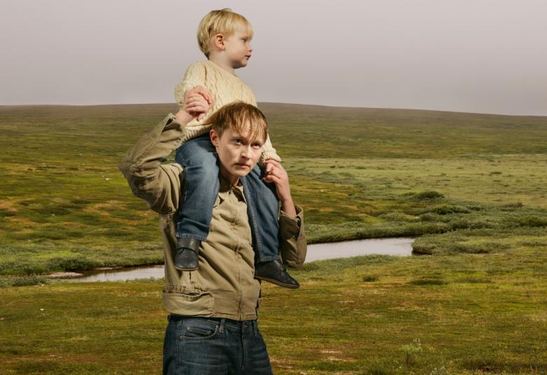 Kuvassa on mies pikkupoika niskassaan ja he ovat kävelemässä tunturimaisessa maastossa. Taustalla on vihreää maastoa ja sinertävän harmaata taivasta.