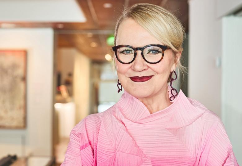 Kuvassa on Anna-Mari Wiljanen puolivartalokuvassa ja hänellä on vaalean punainen pusero päällään. Taustalla näkyy vasemmalla taulu, jossa on vaaleat sävyt.