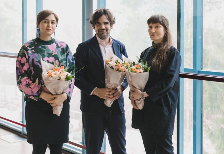 Kuvassa on Bita Razavi vasemmalla isokukallisessa puserossa ja mustassa puolihameessa. Hänen vieressään on mustassa takissa Axel Straschnoy ja oikealla on Johanna Lonka mustissa vaatteissa. Kaikilla on kukat kädessään ja he seisovat isojen ikkunoiden edessä. 