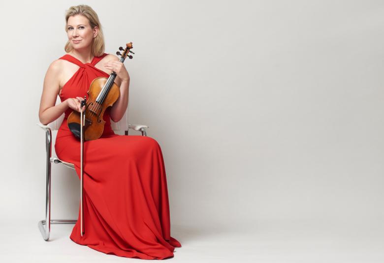 Kuvassa on Elina Vähälä istumassa tuolilla viulu kädessään ja hänellä on punainen puku päällään.  Kuvan tausta on vaalean harmaa.