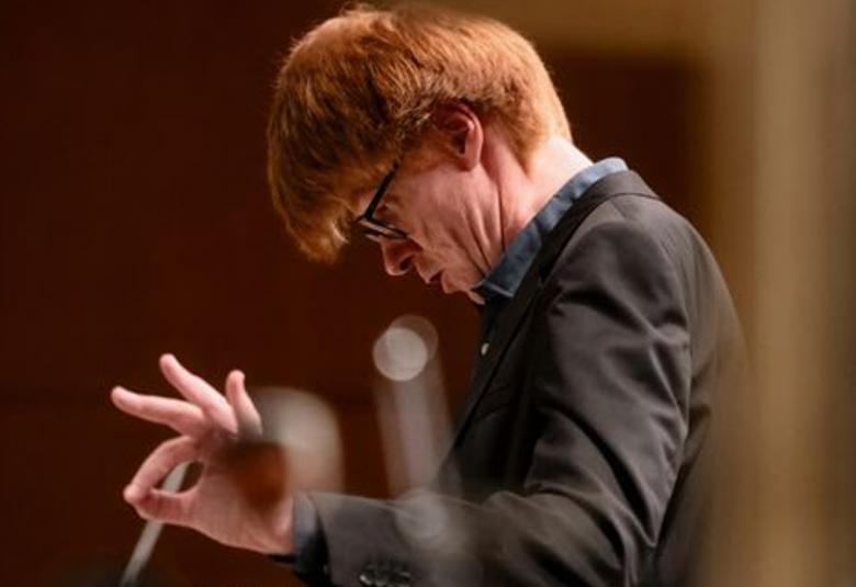 Kuvassa on Enno Poppe sivuttain puolivartalokuvassa johtamassa orkesteria pää alaspäin vasen kämmen sivussa ja sen takaa näkyy tahtipuikko toisessa kädessä. Hänellä on musta pusero ja punaiset hiukset.